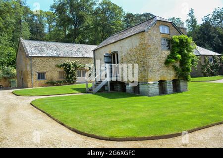 Eine ehemalige Getreidehütte ist heute ein luxuriöses Schlafzimmer auf dem Gelände des neuen Newt Hotels in Somerset. Korn wurde im Zimmer gelagert und getrocknet. Das Gebäude dahinter hat auch Schlafzimmer. Goldene Cotswold-Steine bilden die Wände, während Rosensträucher entlang ihnen eine schöne Szene schaffen. Der Rasen und der Weg sind gut gepflegt und glänzen im Sonnenschein. Stockfoto