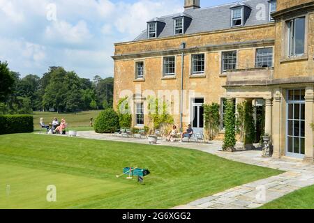 Die Bewohner sonnen sich auf dem Gelände des neuen schönen Newt Hotels in Somerset. Der goldene Cotswold-Stein bildet die Wände, während der Sonnenschein die Szene bedeckt. Der Rasen und der Weg sind gut gepflegt und glänzen im Sonnenschein. Das Gebäude heißt Hadspen House und ist ein restauriertes englisches Landhaus. Stockfoto