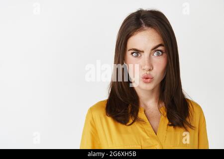 Nahaufnahme einer verwirrten und faszinierten jungen Frau, die eine Augenbraue hochhebt, Lippen stottert und verwirrt auf die Kamera starrt, die auf weißem Hintergrund steht Stockfoto