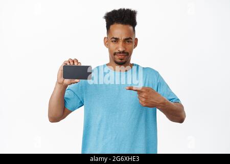 Urteilender afroamerikanischer Mann furche Augenbrauen, zeigt mit dem Finger auf den Smartphone-Bildschirm mit Missbilligung, mag App nicht, schlechte Interface-Anwendung Stockfoto