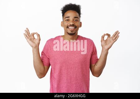 Zen und Geduld. Lächelnder, entspannter afroamerikanischer Mann, der friedlich aussagt, Hände in Zen hält, Yoga-Meditationsgeste, Ruhe bewahren, rosa T tragen Stockfoto