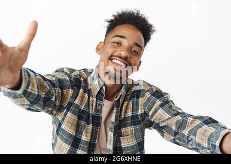 Porträt eines gutaussehenden afroamerikanischen Mannes, fröhlich lächelnd, Hände zur Umarmung spreizen, Freund kuscheln, nach warmer Umarmung greifen, im karierten Hemd stehend Stockfoto