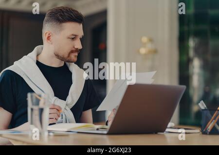 Smart Handsome Mann prüft Informationen auf Papieren funktioniert Remote sitzt zu Hause Büro verwendet modernen Laptop-Computer schreibt in Notebook gekleidet Stockfoto