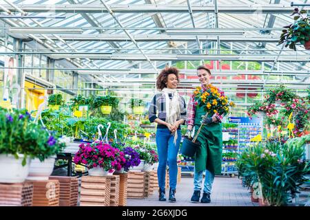 Die ganze Länge einer hübschen und freundlichen Arbeiterin, die einer Kundin beim Kauf einer orangen dekorativen Zimmerpflanze in einem modernen Blumenladen hilft Stockfoto