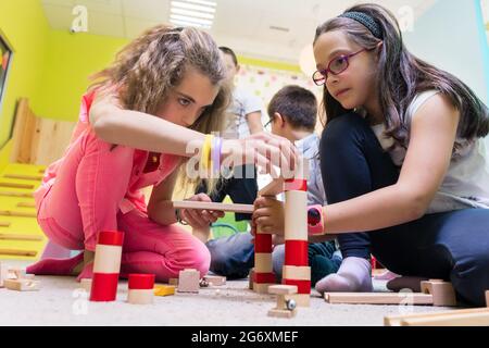 Zwei Pre-school Mädchen zusammen spielen mit Holzspielzeug Bausteine auf dem Boden während der Spielzeit betreut durch eine sorgfältige junge Kindergärtnerin Stockfoto