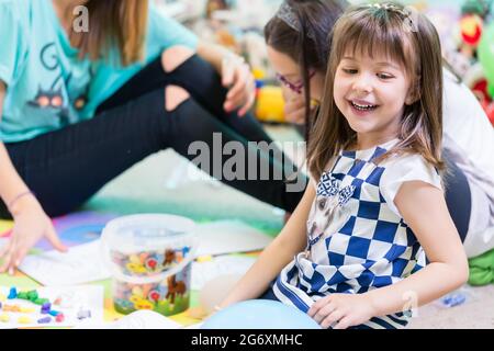 Fröhliche Pre-school Mädchen mit einem trendigen T-Shirt beim Spielen auf dem Fußboden, betreut durch ihre junge und coole Lehrerin in einem modernen Kindergarten Stockfoto