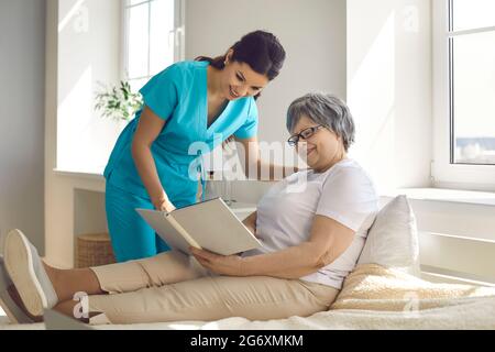 Caregiver in einem Pflegeheim diskutiert ein Buch, das eine ältere Frau liest, während sie im Bett liegt. Stockfoto