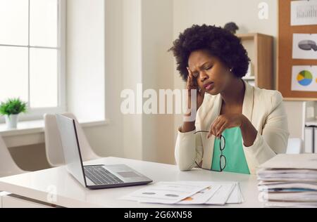 Müde junge Frau mit dunkler Haut, die starke Kopfschmerzen hat und bei der Arbeit vor dem Laptop sitzt. Stockfoto