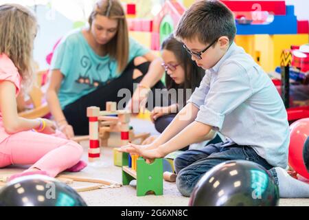 Netter Vorschuljunge, der mit seinen Kollegen beim Bau einer Struktur aus hölzernen Spielzeugblöcken unter der Anleitung eines jungen Kinders zusammenarbeitet Stockfoto