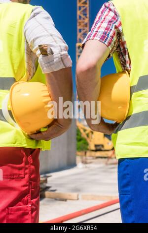 Nahaufnahme der Hände von zwei Arbeitern, die reflektierende Schutzwesten tragen, während sie im Freien auf der Baustelle gelbe Schutzhüte halten Stockfoto