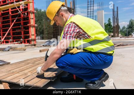 Seitenansicht eines jungen Arbeiters, der eine Sicherheitsweste und einen gelben Harthut trägt, während er einen Nagel in Holz eingeschlagen hat, während der Arbeiten auf der Baustelle eines Contes Stockfoto