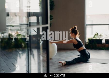 Junge rothaarige schlanke Frau, die mit einem Fitball im Garn sitzt und Stretching-Übungen macht Stockfoto