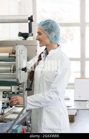Glückliche weibliche Produktionsbetreiberin mit weißem Labormantel und schützender Kopfbedeckung, während sie in einer Kosmetikfabrik mit modernen Geräten arbeitet Stockfoto