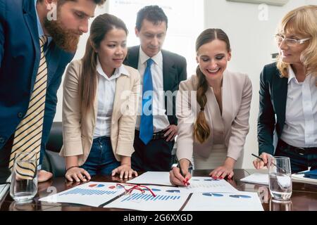 Lächelnde Geschäftsexpertin, die Informationen aus gedruckten Charts während eines interaktiven Meetings mit dem Vorstand im of interpretiert Stockfoto