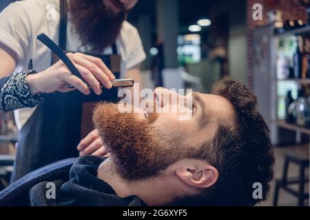 Seitenansicht Kopfaufnahme eines rothaarigen bärtigen jungen Mannes, der lächelt und bereit ist, sich im Friseursalon zu rasieren, eines erfahrenen Barbiers mit einem klassischen Rasiermesser in Hi Stockfoto