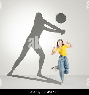 Konzeptuelles Bild mit Mädchen im Vorschulalter und Schatten einer Volleyballspielerin auf hellgrauem Hintergrund. Kindheit und Traum von der großen und berühmten Zukunft. Stockfoto