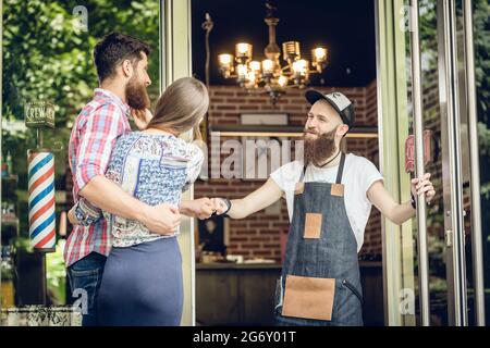 Ein cooler, männlicher Friseur, der einem glücklichen Kunden, einem jungen Mann in Begleitung seiner Freundin, an der offenen Tür eines trendigen Schönheitssalons einen Fauststoß gibt Stockfoto