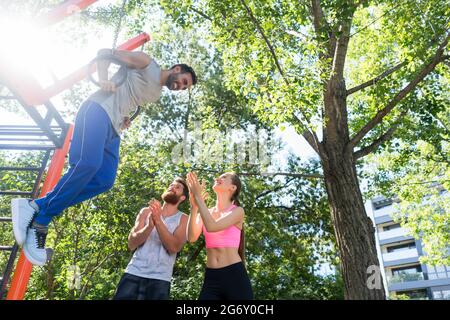 Low-Angle-Ansicht eines starken jungen Mannes, der während des von seinem Freund motivierten Street-Workout-Routings Trizeps an Gymnastikringen schwierig trainiert Stockfoto