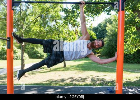 Die ganze Länge eines muskulösen und entschlossenen jungen Mannes lächelt, während er Bodyweight-Übungen in einem modernen Fitnesspark im Sommer macht Stockfoto