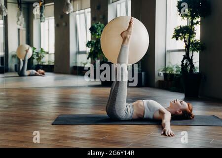 Junge Frau macht Übungen für Beine mit Pilates Ball, während sie auf Yoga-Matte liegt Stockfoto