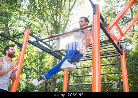 Low-Angle-Ansicht eines starken und entschlossenen jungen Mannes, der während des extremen Outdoor-Workouts in den Applaus seiner Freunde erstaunliche Pull-up-Variationen macht Stockfoto