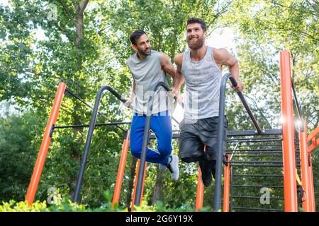 Blick aus der Tiefe auf zwei hübsche junge Männer, die sich leidenschaftlich für Fitness begeistern und in einem Calisthenics Park mit modernen Geräten Trizeps trainieren Stockfoto