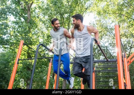 Blick aus der Tiefe auf zwei hübsche junge Männer, die sich leidenschaftlich für Fitness begeistern und in einem Calisthenics Park mit modernen Geräten Trizeps trainieren Stockfoto