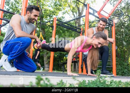 Eine tief anliegende Ansicht einer jungen Frau, die eine Beinverlängerung trainiert, mit einem Suspension Trainer, der von ihren fröhlichen, sportlichen Freunden in einem modernen calis motiviert wurde Stockfoto