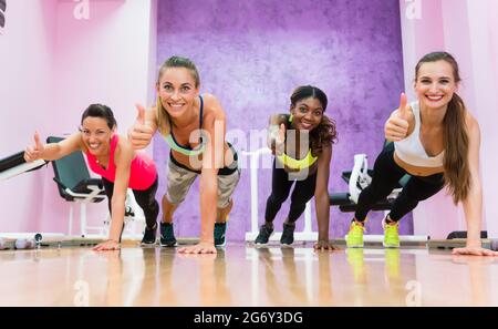 Gruppenportrait von vier glücklichen Frauen, die von der Grundposition der Planke den Daumen nach oben zeigen, als gleiches Zeichen für einen effizienten Trainingskurs in einem trendigen Fitnessclub Stockfoto