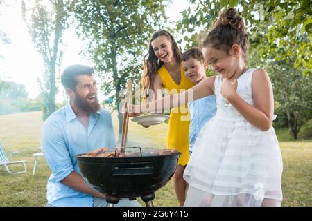 Nettes lustiges Mädchen mit Holzzangen während der Zubereitung von Fleisch auf dem Grill Holzkohlegrill vor ihrem Vater, Mutter und Bruder während des Picknicks im Sommer Stockfoto
