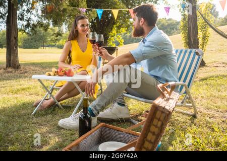Junge und schöne Paar in Liebe Toasten mit Rotwein, während Sie sitzen auf Klappstühlen beim romantischen Picknick im Grünen Stockfoto