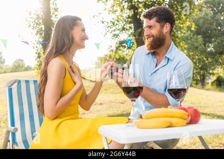 Schöne junge Frau, die mit Freude und Emotion den Heiratsantrag ihres Freundes während eines romantischen Picknicks an einem sonnigen Sommertag annimmt Stockfoto