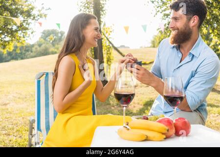 Schöne junge Frau, die mit Freude und Emotion den Heiratsantrag ihres Freundes während eines romantischen Picknicks an einem sonnigen Sommertag annimmt Stockfoto