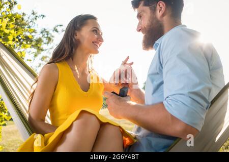 Aus der Perspektive eines glücklichen jungen Mannes, der einen Verlobungsring auf den Finger seiner Freundin legt, nachdem er seinen Vorschlag angenommen hatte, während er zusammen saß Stockfoto