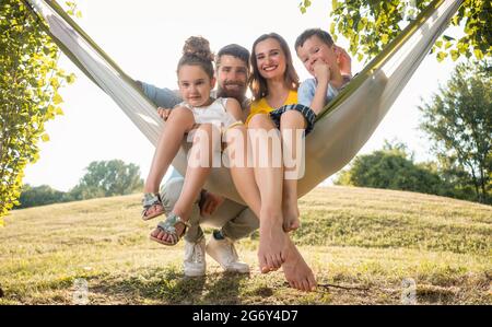 Family Portrait mit einer schönen Mutter von zwei Verspielte Kinder schwingen in der Hängematte, während bei camera Suchen neben ihrem Ehemann im Freien im Sommer Stockfoto