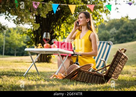 Schöne und aktive junge Frau, die mit dem Handy und Tablet spricht, während sie auf einem Klappstuhl sitzt und ein entspannendes Picknick im Park macht Stockfoto