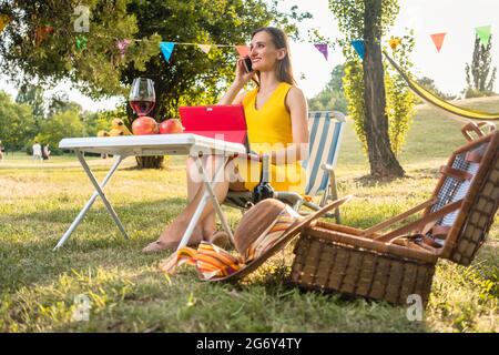 Schöne und aktive junge Frau, die mit dem Handy und Tablet spricht, während sie auf einem Klappstuhl sitzt und ein entspannendes Picknick im Park macht Stockfoto