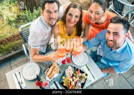 Ein Porträt aus dem Blickwinkel von vier fröhlichen und schönen jungen Freunden, die in einem trendigen Restaurant mit köstlichen Speisen in die Kamera schauen und dabei die Kamera anstoßen Stockfoto