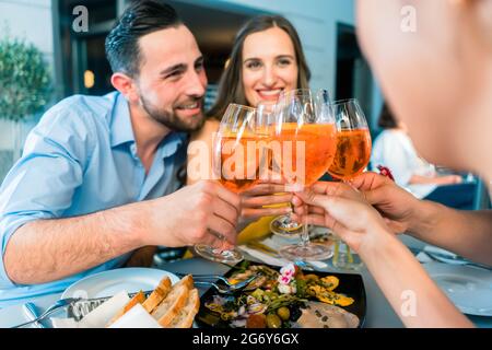 Hübscher junger Mann aus dem Nahen Osten, der neben seiner kaukasischen Freundin sitzt, während er mit zwei gemeinsamen Freunden in einem trendigen Restaurant in s im Freien toaste Stockfoto