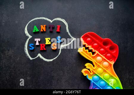 Toy Pop it Dinosaurier Regenbogenfarben auf schwarzem Hintergrund mit bunten Buchstaben und der Aufschrift-Anti-Stress in einer Sprechblase. Stockfoto