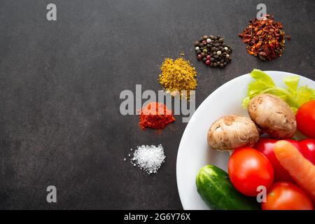 Ein weißer Teller mit frischem Gemüse: Pilze, Tomaten, Karotten und Kräutern, um die verschiedene Gewürze in Haufen verstreut sind. Auf schwarzem Hintergrund Stockfoto