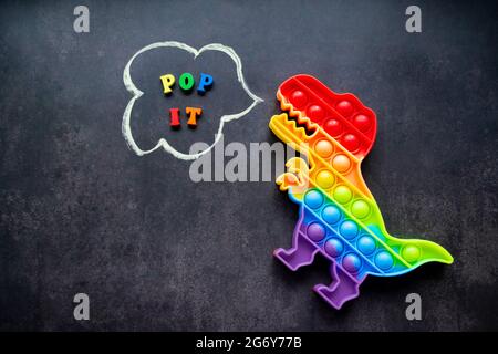 Pop it Dinosaurier Spielzeug Regenbogenfarben auf schwarzem Hintergrund mit bunten Buchstaben und der Inschrift - Pop it in einer Sprechblase. Stockfoto