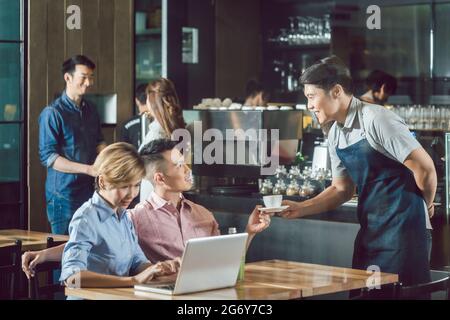 Fröhlicher Kellner, der männlichen Gästen Kaffee serviert, die neben einer Frau sitzen, die einen Laptop benutzt Stockfoto