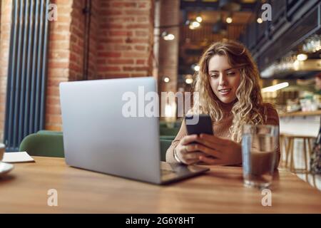 Frau, die Smartphone benutzt, während sie mit einem Laptop im Café sitzt Stockfoto