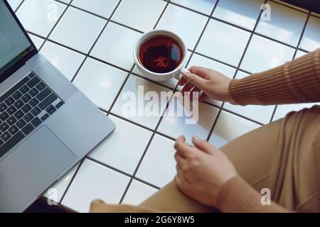 Frau, die neben dem Fenster mit einer Tasse Tee und einem Laptop sitzt Stockfoto