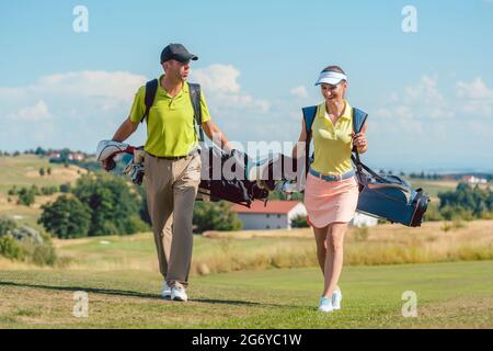 Volle Länge des ein glückliches Paar mit einem gesunden Lebensstil golf Outfits tragen, während die Ständer Taschen mit professionellen Vereine in Richtung Golfplatz Stockfoto