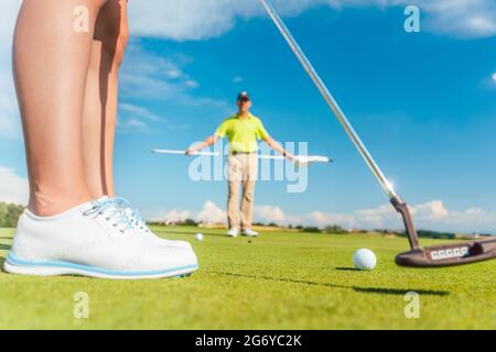 Golfball auf dem Putting Green im selektiven Fokus hinter dem unteren Teil einer weiblichen Spielerin während der Klasse, mit einem erfahrenen Lehrer auf professionelle Stockfoto