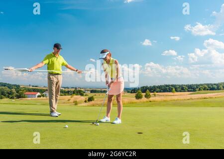 Die ganze Länge einer Frau, die die Flugbahn des Balls zum Loch berechnet, während sie mit ihrem männlichen Partner oder Instruktor beim Profigolf spielt Stockfoto