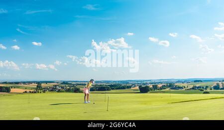 Die ganze Länge einer entschlossenen jungen Frau, die Golf übt, bewegt sich und schlägt auf das Gras eines Trainingsgebietes zum Putting Green, in einem idyllischen sonnigen ländlichen Gebiet Stockfoto