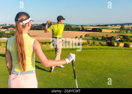 Rückansicht eines professionellen Golfers, der in der Zielposition eines langen Schusses steht, während eines schwierigen Matches mit seiner Partnerin in einem sonnigen d Stockfoto